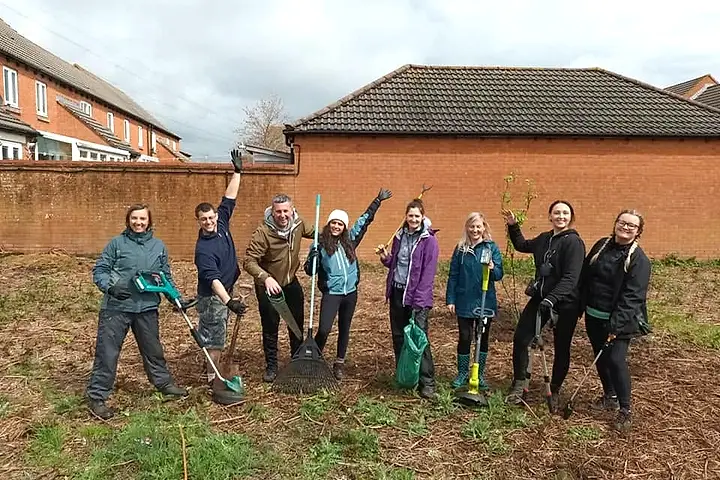 Eight smiling volunteers with rakes, strimmers and litter pickers on a freshly cleared patch of ground beside a brick wall
